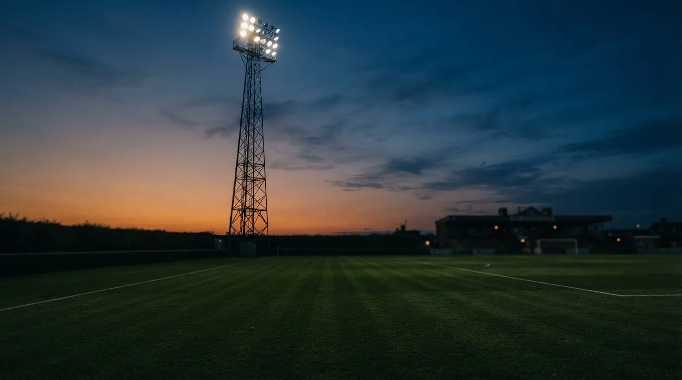 Ein Flutlichtmast ragt in den Abendhimmel, die Rasenfläche darunter liegt leer und dunkelgrün da.