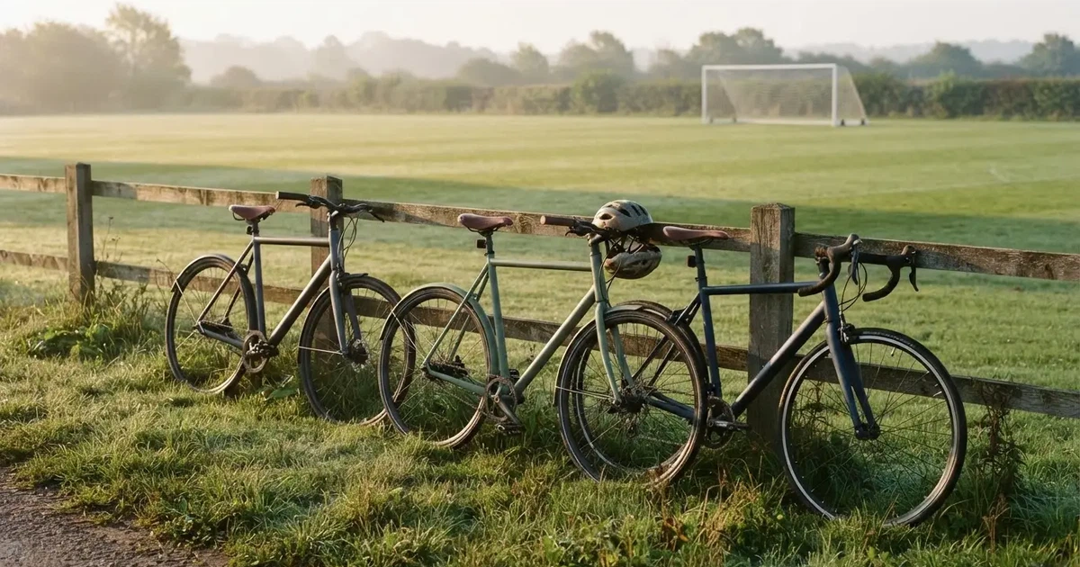 Drei Fahrräder lehnen nebeneinander an einem Zaun vor einem Sportplatz, dahinter ein grüner Rasen im Dunst.
