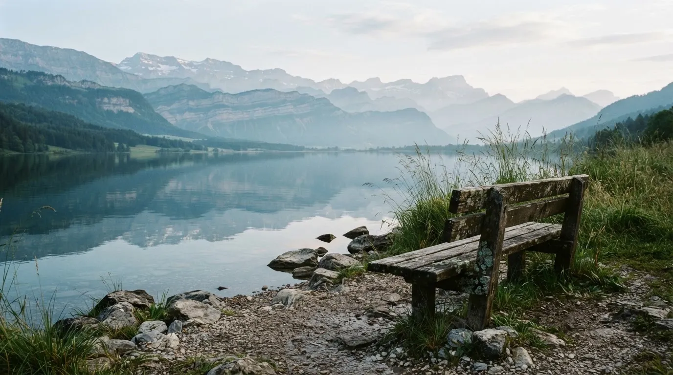 Eine Holzbank an einem Seeufer mit Blick auf eine Bergkette im Morgendunst, das Wasser ist spiegelglatt.