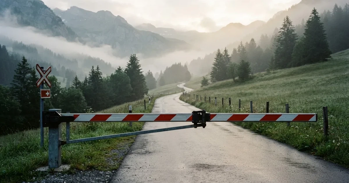 Eine geschlossene Schranke steht an einer ruhigen Bergstrasse im Morgennebel, dahinter Tannen und eine nasse Wiese.